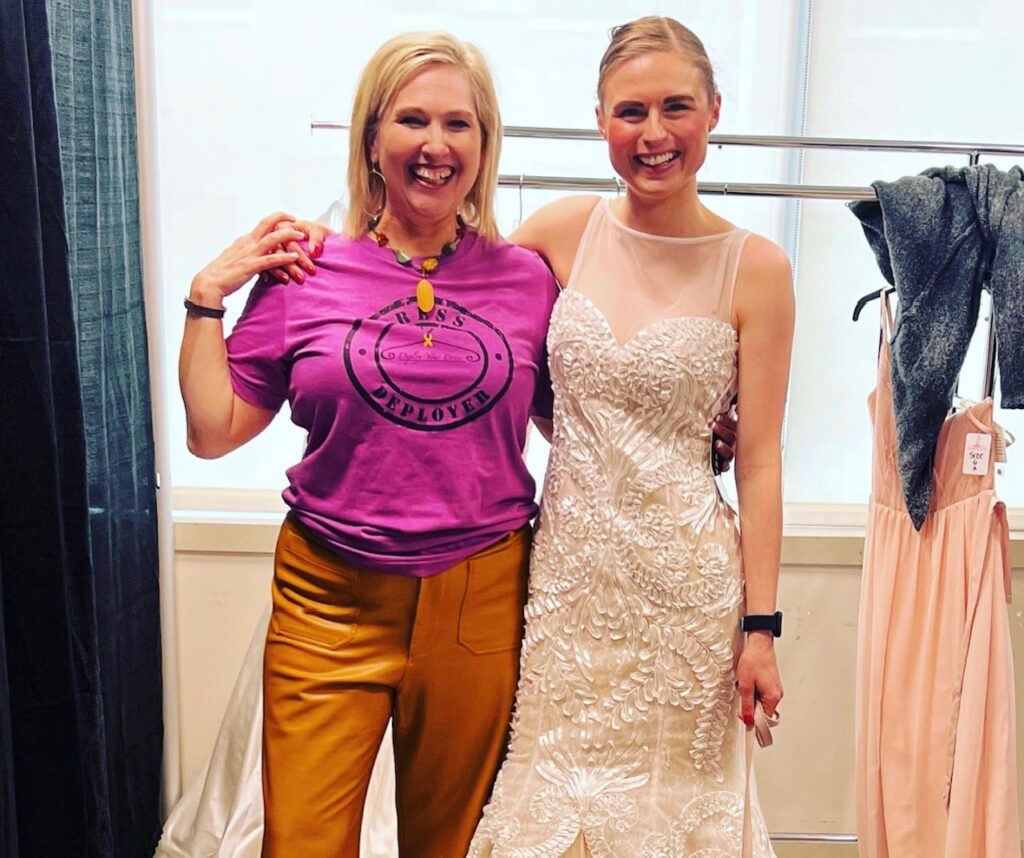 Two women posing for a picture in front of some dresses.