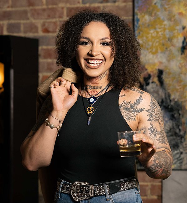 A woman with tattoos holding a glass of whiskey.